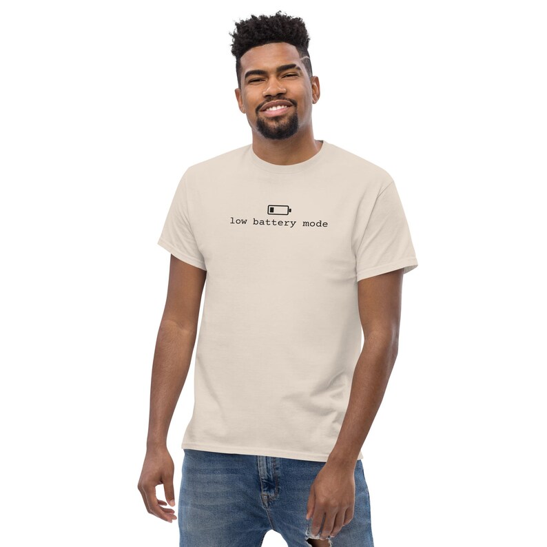 May include: A beige short-sleeved t-shirt featuring a black graphic of a battery icon and the text "low battery mode" printed in a typewriter-style font across the center of the chest. The shirt has a crew neckline and a standard fit.
