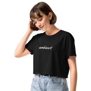 May include: A black cropped t-shirt featuring the word ambivert written in a white cursive script font across the center of the chest. The garment has short sleeves and a round neckline, paired with light blue denim jeans with a visible button.