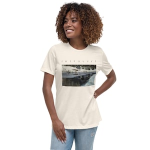 May include: A cream-colored short-sleeve t-shirt featuring the word "introvert" printed in black lowercase letters above a rectangular graphic depicting a snowy landscape with a frozen river and dark trees under a winter sky.