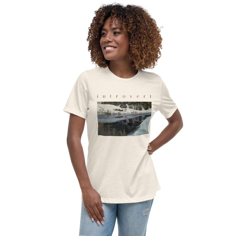 May include: A cream-colored short-sleeve t-shirt featuring the word "introvert" printed in black lowercase letters above a rectangular graphic depicting a snowy landscape with a frozen river and dark trees under a winter sky.