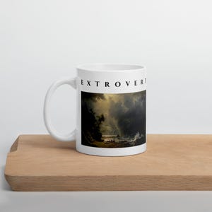 May include: A white ceramic mug featuring the word EXTROVERT printed in black capital letters above a reproduction of a dark, dramatic landscape painting depicting a stormy coastal scene with turbulent clouds, crashing waves, and rocky cliffs.