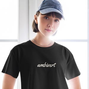 May include: A black short-sleeved t-shirt featuring the word ambivert printed in a white cursive script font across the chest. The shirt has a classic crew neckline and a standard fit, providing a clean and simple look for casual daily wear.
