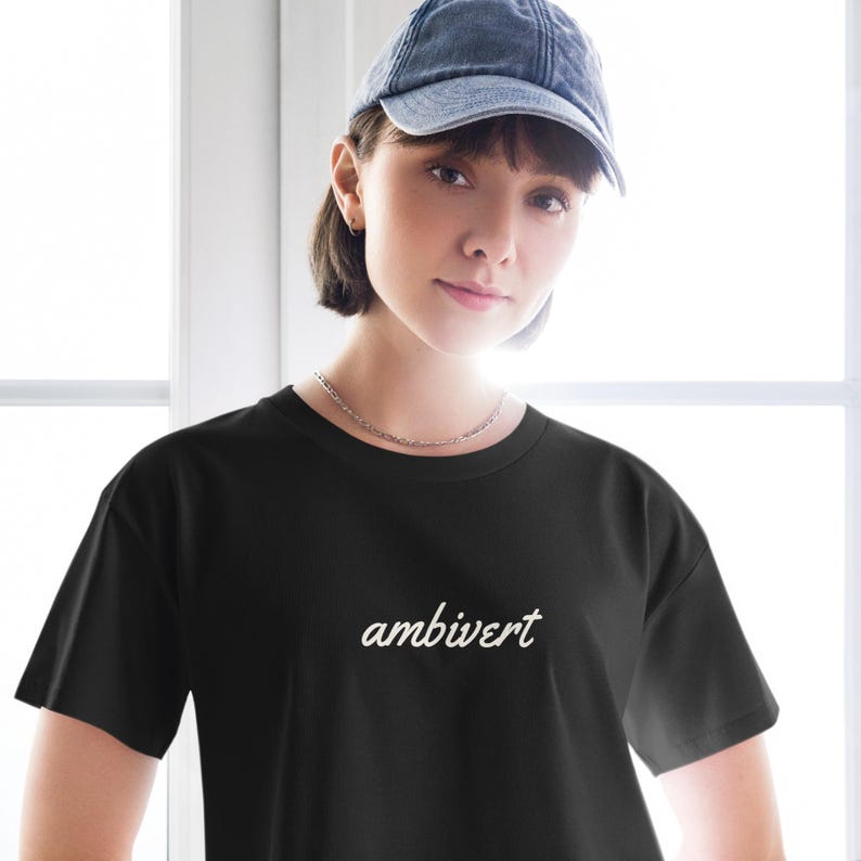 May include: A black short-sleeved t-shirt featuring the word ambivert printed in a white cursive script font across the chest. The shirt has a classic crew neckline and a standard fit, providing a clean and simple look for casual daily wear.