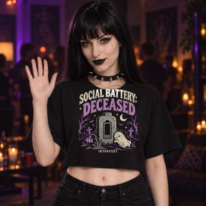 May include: A black cropped t-shirt featuring a graphic print with the text "SOCIAL BATTERY: DECEASED" and "INTROVERT". The design includes a tombstone shaped like a battery, a small ghost, a crescent moon, bare trees, and purple flowers.