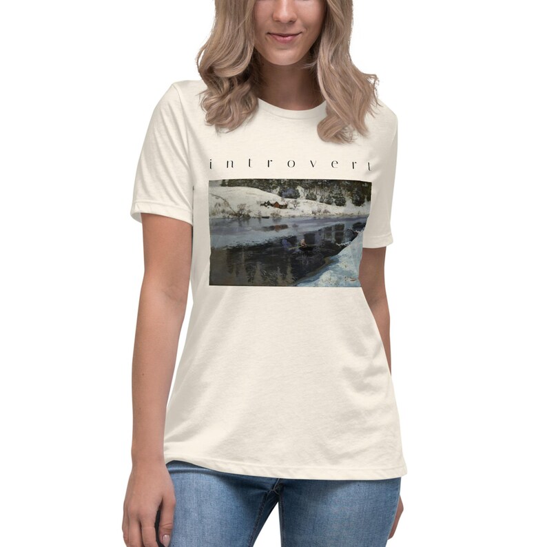 May include: A cream-colored short-sleeve t-shirt featuring the word "introvert" printed in black text above a rectangular graphic. The graphic depicts a snowy landscape with a dark, reflective body of water surrounded by white, snow-covered banks.