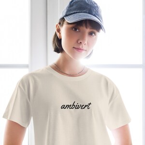 May include: A cream-colored short-sleeve t-shirt featuring the word ambivert printed in a black cursive script font across the chest. The garment has a crew neckline and a standard fit, providing a clean and simple aesthetic for casual wear.