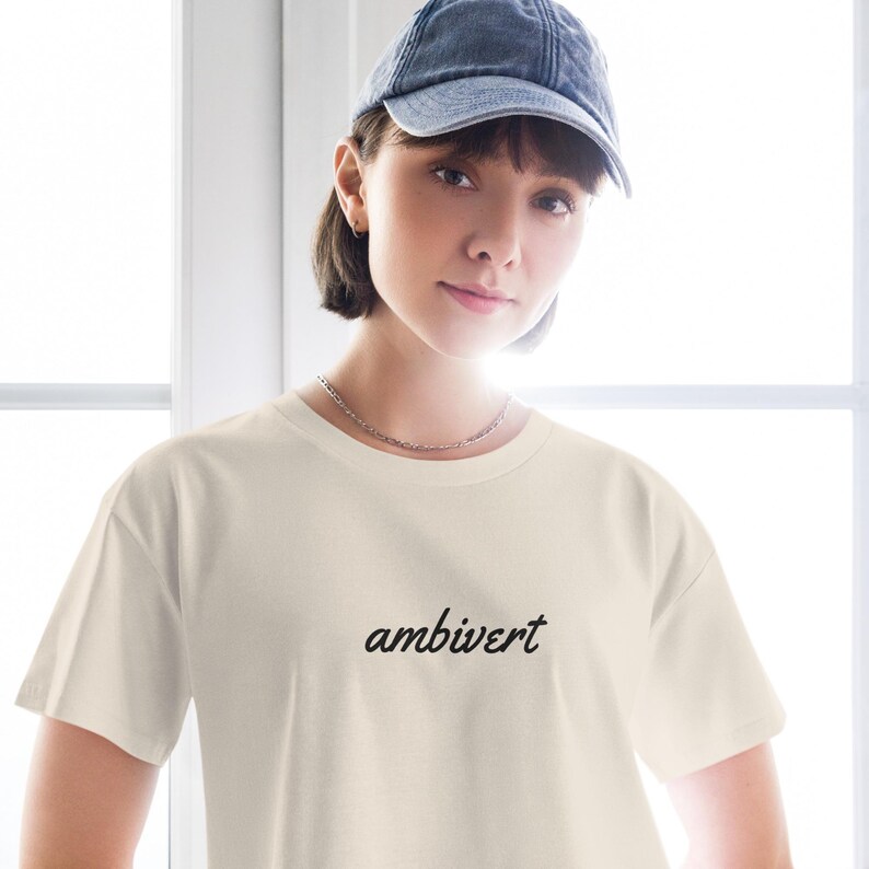 May include: A cream-colored short-sleeve t-shirt featuring the word ambivert printed in a black cursive script font across the chest. The garment has a crew neckline and a standard fit, providing a clean and simple aesthetic for casual wear.
