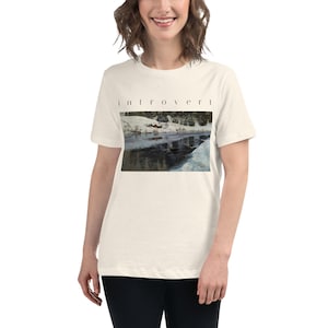 May include: A cream-colored short-sleeve t-shirt featuring the word "introvert" printed in black lowercase letters above a rectangular graphic depicting a snowy landscape with a dark, reflective body of water and surrounding trees.
