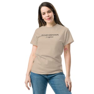 May include: A beige short-sleeved t-shirt featuring the black printed text fellow extrovert we approve on the chest. The garment has a crew neckline and a standard fit, paired with blue denim jeans that have a classic five-pocket design.