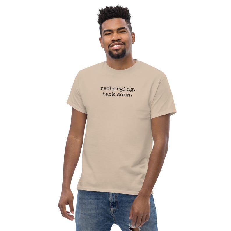 May include: A beige short-sleeved t-shirt featuring the black text "recharging. back soon." printed in a typewriter-style font across the center of the chest. The garment has a crew neckline and a standard fit, paired with blue denim jeans.