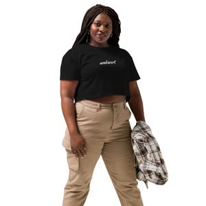 May include: A black short-sleeved crop top featuring the word ambivert written in white cursive script across the chest. The garment is paired with tan cargo pants and a plaid button-down shirt held in one hand, completing the casual outfit.