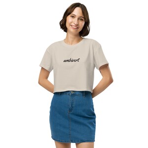 May include: A beige cropped t-shirt featuring the word ambivert printed in a black cursive script across the chest. The garment has short sleeves and a crew neckline, paired with a blue denim skirt that has a button and zipper closure.