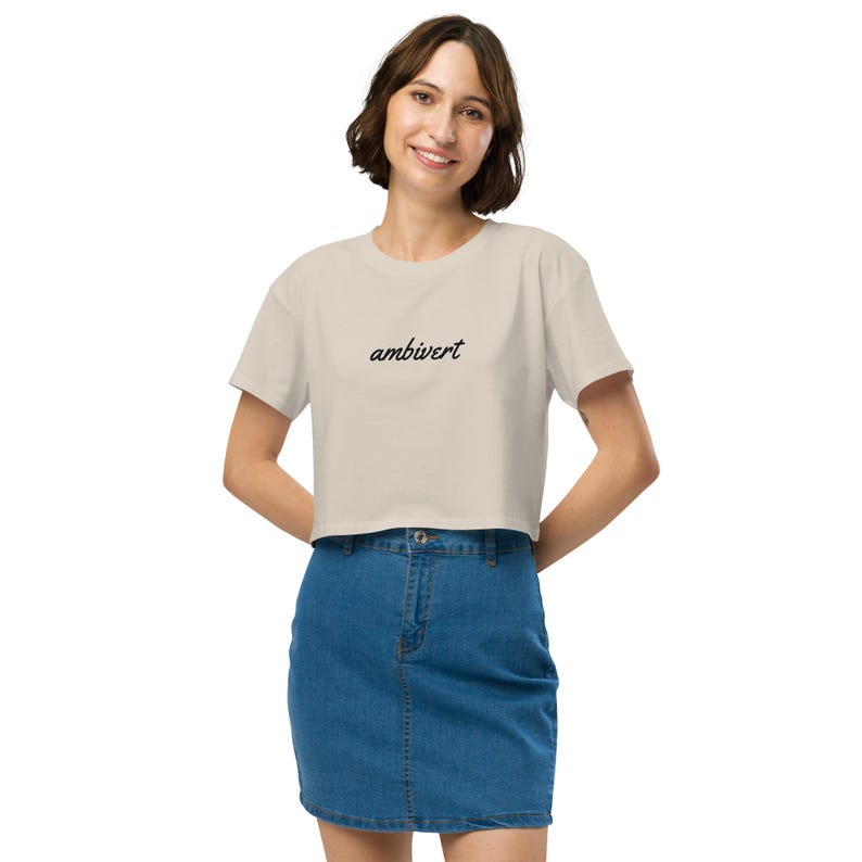 May include: A beige cropped t-shirt featuring the word ambivert printed in a black cursive script across the chest. The garment has short sleeves and a crew neckline, paired with a blue denim skirt that has a button and zipper closure.