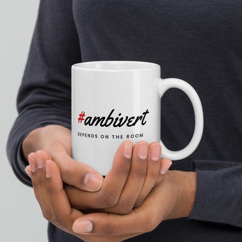 May include: A white ceramic mug featuring the red hashtag symbol followed by the black text #ambivert and the black text DEPENDS ON THE ROOM printed in a sans-serif font below it. The mug has a standard handle and a smooth, glossy finish.