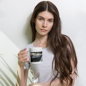 Puede incluir: Una taza de cer&aacute;mica blanca con una imagen impresa de un bosque nevado reflejado en un cuerpo de agua oscuro. La palabra "introvert" est&aacute; impresa en letras may&uacute;sculas peque&ntilde;as, negras y sin remates sobre el gr&aacute;fico del paisaje.