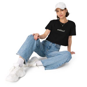 May include: A black short-sleeved crop top featuring the word ambivert printed in white cursive script across the chest, paired with light blue denim jeans, white socks, white sneakers, and a plain white baseball cap worn on the head.