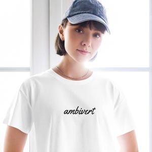May include: A white short-sleeved t-shirt featuring the word ambivert printed in a black cursive script across the chest. The garment is paired with a dark grey denim baseball cap and a thin silver chain necklace worn around the neck.