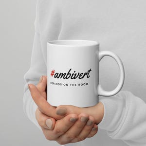 May include: A white ceramic mug featuring the red hashtag symbol followed by the black cursive text #ambivert and the black sans-serif text DEPENDS ON THE ROOM printed on the front surface of the mug, which has a standard handle.