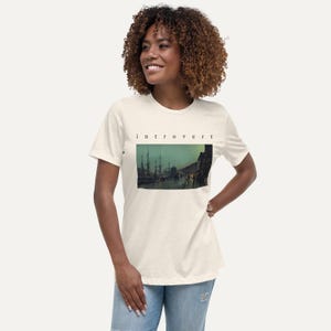 May include: A cream-colored short-sleeve t-shirt featuring the word "introvert" printed in black lowercase letters above a rectangular graphic depicting a dark, moody harbor scene with tall ship masts and silhouettes of buildings at twilight.