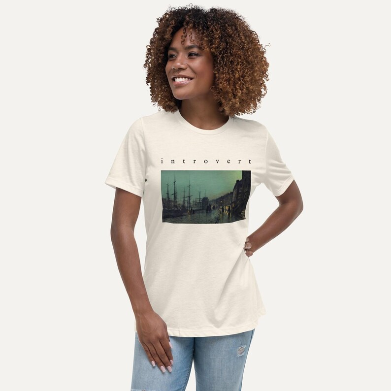 May include: A cream-colored short-sleeve t-shirt featuring the word "introvert" printed in black lowercase letters above a rectangular graphic depicting a dark, moody harbor scene with tall ship masts and silhouettes of buildings at twilight.