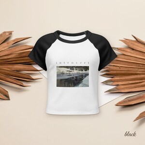 May include: A white raglan t-shirt with black short sleeves and a black crew neckline. The center of the shirt features a rectangular graphic print of a snowy landscape with a body of water and the word introvert printed in black text above it.