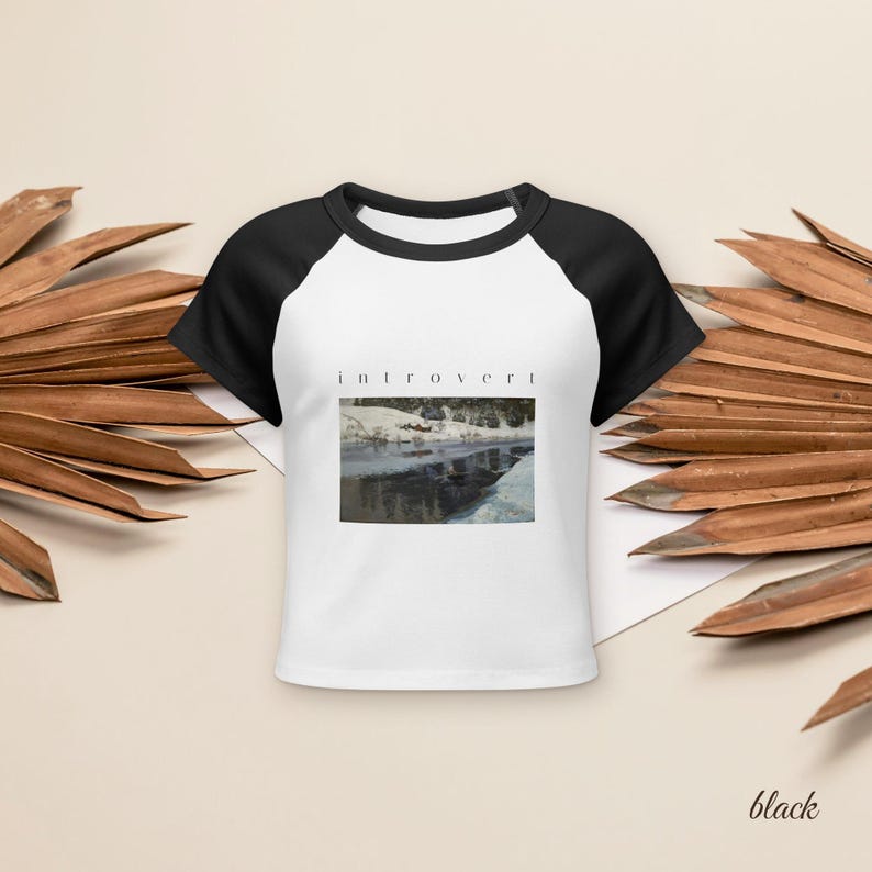 May include: A white raglan t-shirt with black short sleeves and a black crew neckline. The center of the shirt features a rectangular graphic print of a snowy landscape with a body of water and the word introvert printed in black text above it.