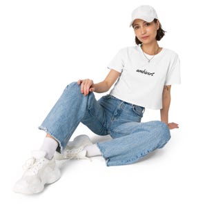 May include: A white baseball cap, a white short-sleeved crop top with the word ambivert printed in black script, blue denim jeans with frayed hems, white socks, and white athletic sneakers with thick soles and layered paneling details.