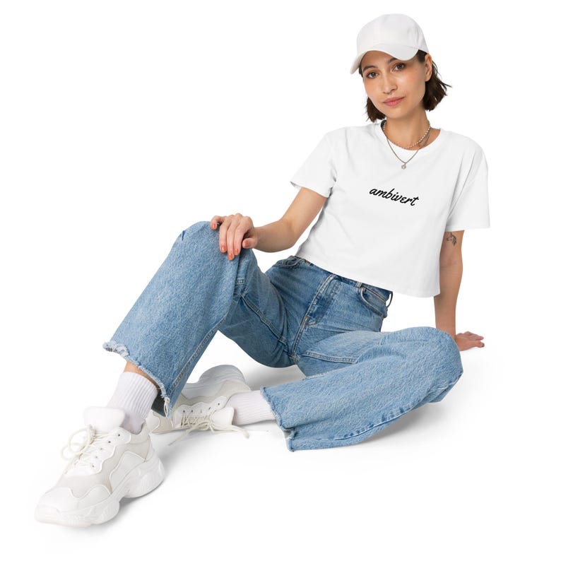 May include: A white baseball cap, a white short-sleeved crop top with the word ambivert printed in black script, blue denim jeans with frayed hems, white socks, and white athletic sneakers with thick soles and layered paneling details.