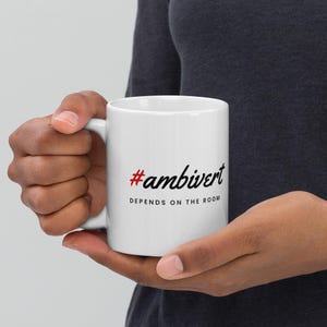 May include: A white ceramic mug featuring the red hashtag symbol followed by the black text #ambivert in a script font, with the phrase DEPENDS ON THE ROOM printed in black capital letters underneath the hashtag on the side of the mug.