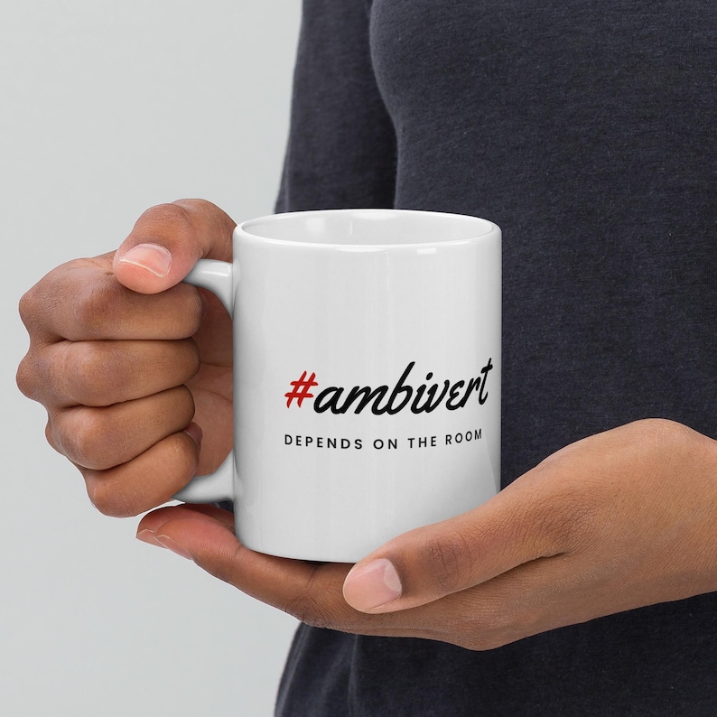 May include: A white ceramic mug featuring the red hashtag symbol followed by the black text #ambivert in a script font, with the phrase DEPENDS ON THE ROOM printed in black capital letters underneath the hashtag on the side of the mug.