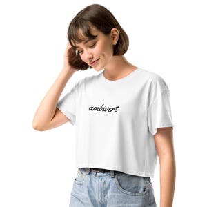 May include: A white cropped t-shirt featuring the word ambivert printed in a black cursive script across the center of the chest. The garment has short sleeves and a crew neckline, providing a simple and clean aesthetic for casual wear.