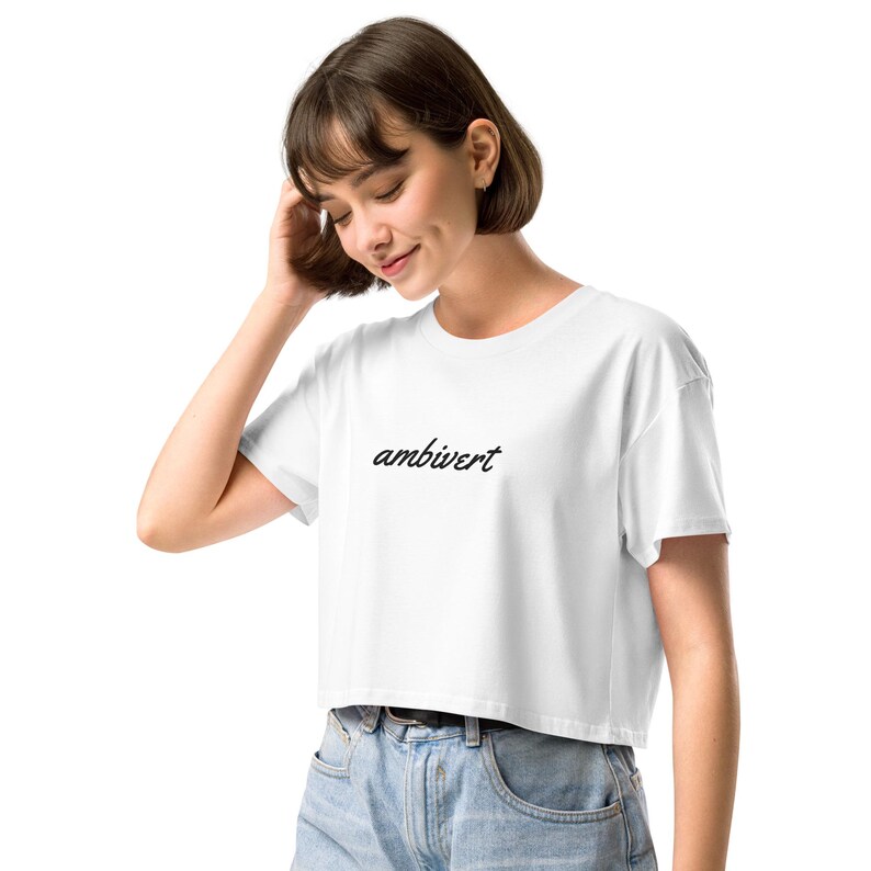 May include: A white cropped t-shirt featuring the word ambivert printed in a black cursive script across the center of the chest. The garment has short sleeves and a crew neckline, providing a simple and clean aesthetic for casual wear.