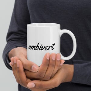 May include: A white ceramic mug featuring the word ambivert printed in a black cursive script font. The mug has a smooth cylindrical body and a curved handle on the right side, providing a simple and clean design for holding hot or cold beverages.