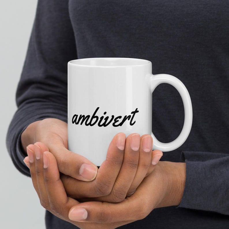 May include: A white ceramic mug featuring the word ambivert printed in a black cursive script font. The mug has a smooth cylindrical body and a curved handle on the right side, providing a simple and clean design for holding hot or cold beverages.