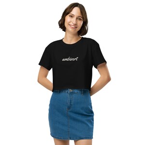 May include: A black short-sleeved crop top featuring the word ambivert written in a white cursive script font across the chest. The garment is paired with a blue denim skirt that has a button and zipper closure at the center front.