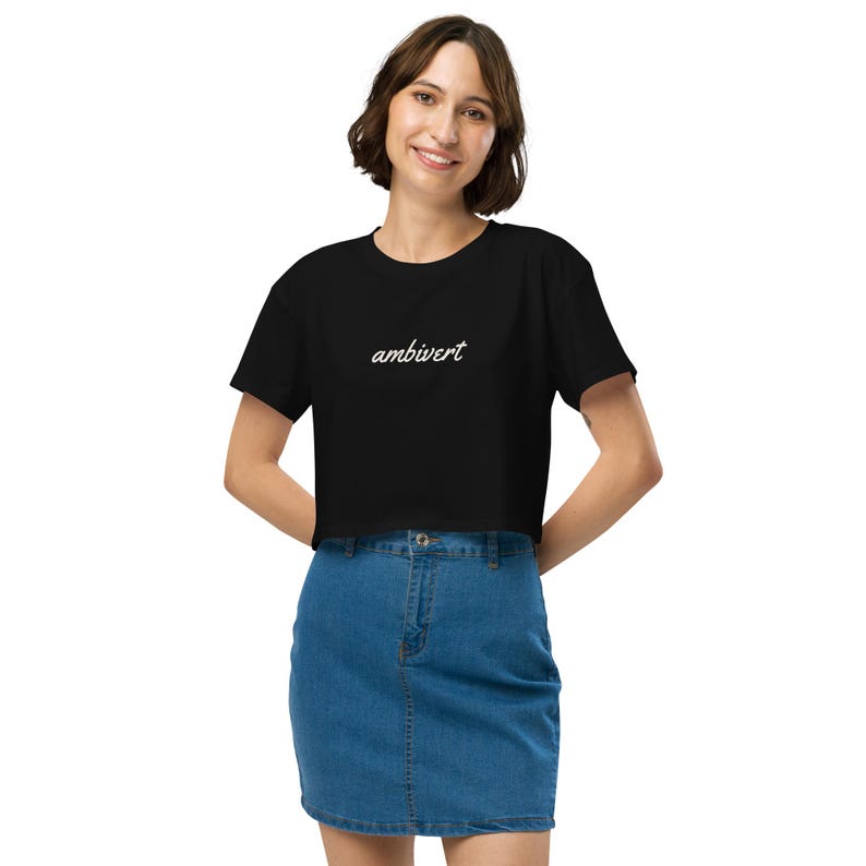 May include: A black short-sleeved crop top featuring the word ambivert written in a white cursive script font across the chest. The garment is paired with a blue denim skirt that has a button and zipper closure at the center front.