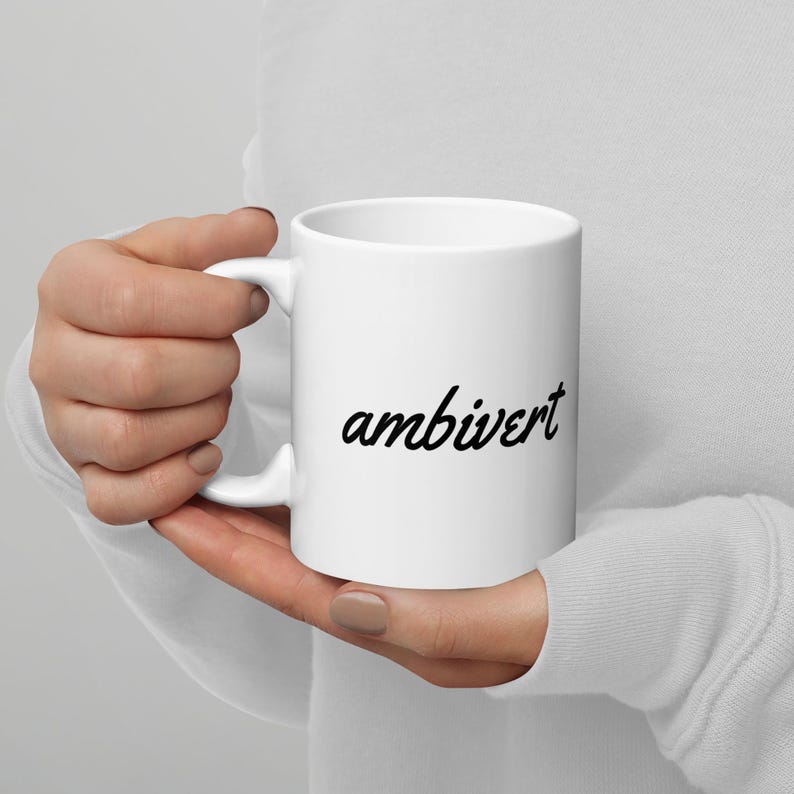 May include: A white ceramic mug featuring the word ambivert printed in a black cursive script font on the front. The mug has a standard cylindrical shape with a curved handle on the side, providing a simple and clean design for daily beverage use.