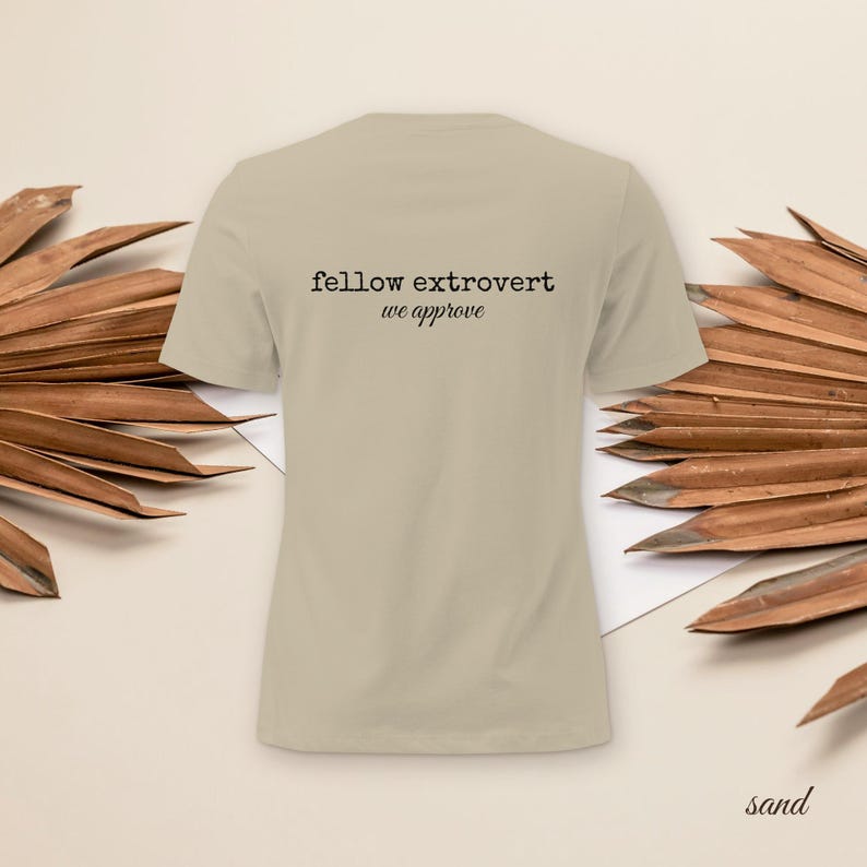 May include: A sand-colored short-sleeve t-shirt featuring the black text "fellow extrovert we approve" printed on the center of the chest. The garment has a crew neckline and a standard fit, presented in a neutral tone labeled as sand.