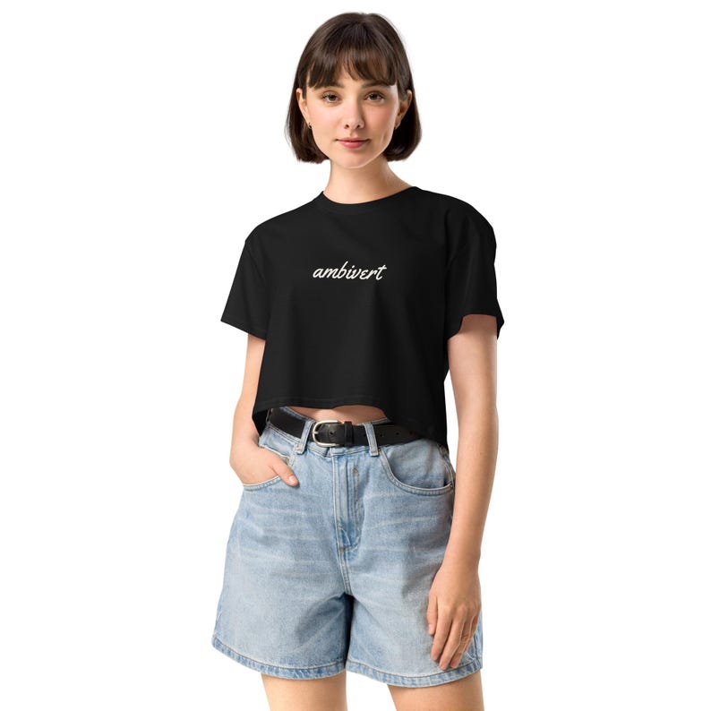 May include: A black cropped t-shirt featuring the word ambivert printed in white cursive script across the chest. The shirt has short sleeves and a crew neckline, paired with light blue denim shorts that have a black belt with a silver buckle.