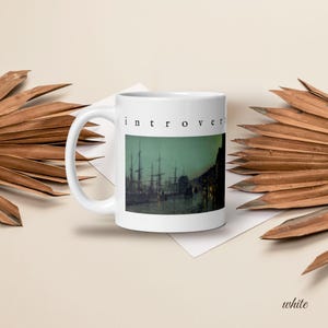May include: A white ceramic mug featuring a printed image of a dark, moody harbor scene with tall sailing ships at dusk. Above the image, the word "introvert" is printed in a simple, black, lowercase serif font. The mug has a curved handle.