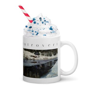 Puede incluir: Una taza de cer&aacute;mica blanca con la palabra introvert impresa en negro sobre una pintura de paisaje invernal de una orilla nevada y una peque&ntilde;a caba&ntilde;a roja. La taza est&aacute; cubierta con nata montada, virutas azules y una pajita a rayas rojas y blancas.