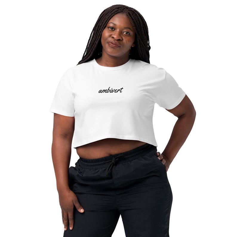 May include: A white short-sleeved crop top featuring the word ambivert embroidered in black cursive script across the center of the chest. The garment has a crew neckline and a straight hemline that sits above the waistline of the black pants.
