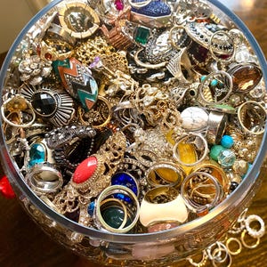 May include: A glass bowl filled with a variety of rings, including gold, silver, and gemstone rings. The rings are of different styles and sizes, and some have intricate designs.