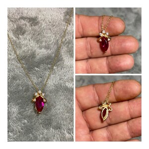 May include: A gold necklace featuring a marquise-cut red gemstone pendant accented with three small clear stones at the top. The pendant is suspended from a delicate gold chain, showcasing a vibrant red center stone and a polished gold setting.