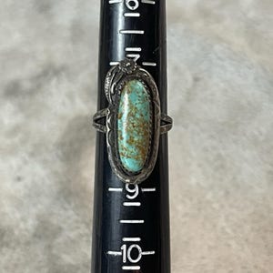 May include: A silver ring featuring an oval turquoise stone with brown matrix, set on a black ring mandrel. The metal setting has decorative etched details and a small flower accent at the top. The ring is positioned near the size 9 mark.
