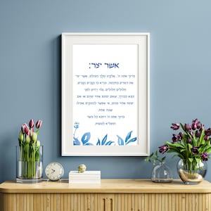 May include: A framed print with a blue and white watercolor design featuring a floral motif. The print includes a Hebrew text that reads "Aser Yitzar" which translates to "Who Created."