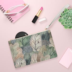 Könnte beinhalten: Eine rechteckige Clutch mit einem Waldmuster in Grün-, Grau- und Beigetönen. Die Clutch hat einen goldenen Reißverschluss und eine weiße Handschlaufe. Weitere Artikel auf der rosa Oberfläche sind Sonnenbrille, Lippenstift und eine Sukkulente.