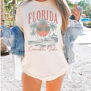 May include: Cream-coloured sweatshirt with a Florida graphic. The design features the word "FLORIDA" in red, palm trees, a sunset, and an alligator. The text "Crocodile State" is below the graphic. The sweatshirt has long sleeves.