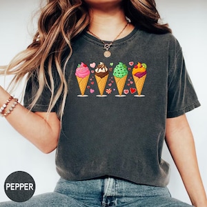 Peut inclure: T-shirt gris foncé orné d'un motif de cônes de crème glacée colorés. Le motif présente quatre cornets de glace aux saveurs et garnitures variées, comme de la glace rose, au chocolat, verte et multicolore. Les cônes sont décorés de cœurs et autres petits détails.