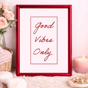Könnte beinhalten: Ein roter Bilderrahmen mit dem Schriftzug "Good Vibes Only" in roter Schrift. Der Rahmen liegt auf einer flauschigen weißen Oberfläche, mit einer Kerze, Blumen und Schmuck in der Nähe. Das Thema ist Entspannung und Positivität.